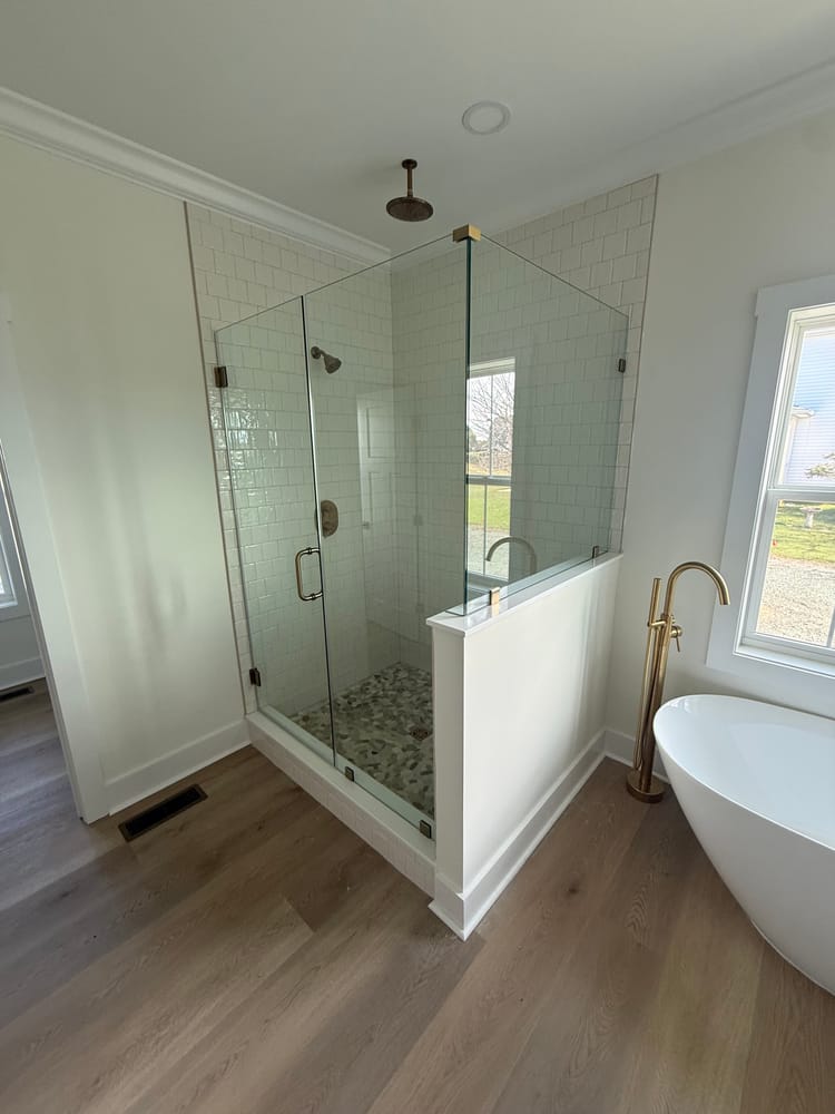 Frameless glass enclosure with brushed brass hinges and handle on a white knee wall, next to a freestanding tub